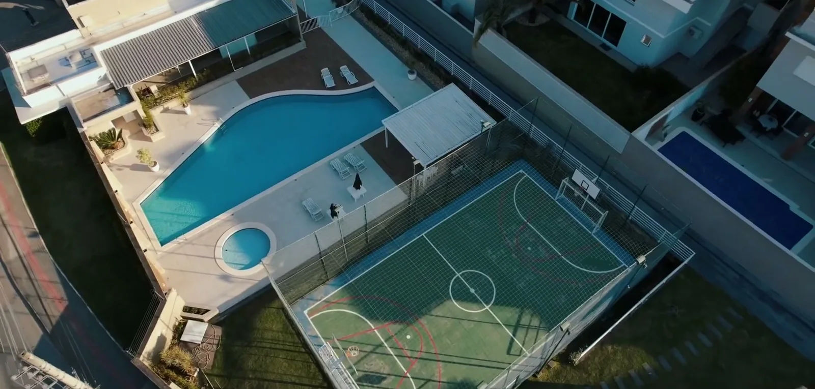 Vista de cima da área de lazer do Parque da Pedra: piscina adulto, piscina infantil redonda, deck com espreguiçadeiras e a quadra poliesportiva cercada