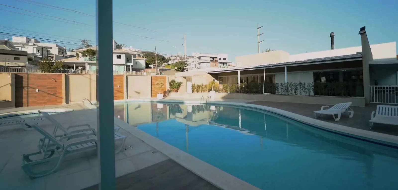 Piscina adulto do condomínio com espreguiçadeiras no deck e o salão de festas ao fundo