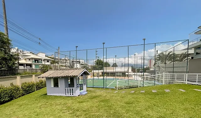 Gramado do condomínio com a casinha de brincar e a quadra cercada ao fundo
