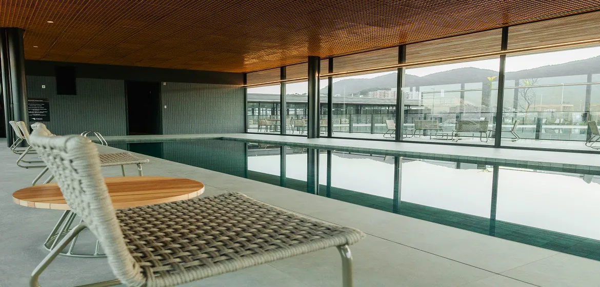 Piscina coberta aquecida do clube, com raia, forro de madeira e parede de vidro para o condomínio